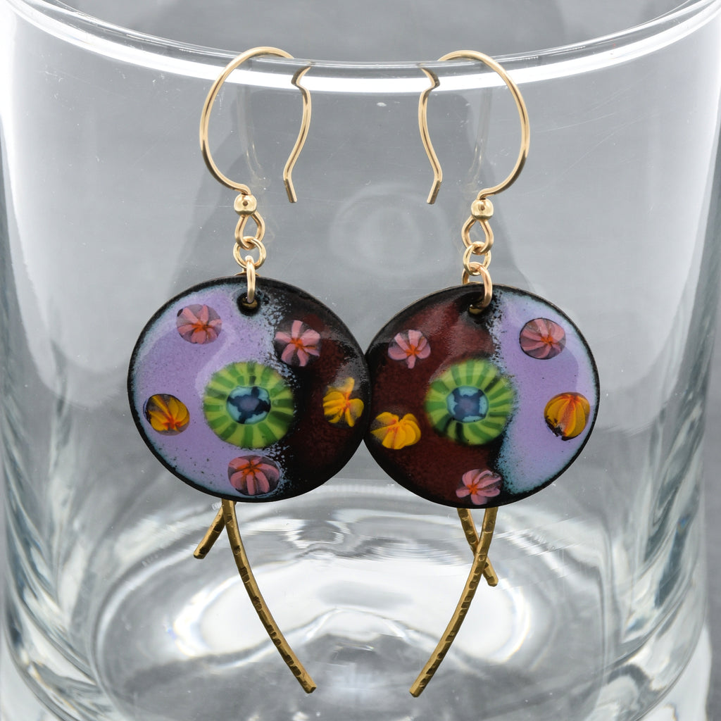 A pair of handmade drop earrings with circular lavender and burgundy enamel charms featuring colorful floral patterns, suspended from gold ear  wires and brass textured daggers, displayed inside a clear glass tumbler - made with love in Colorado by Ruby Acorn.
