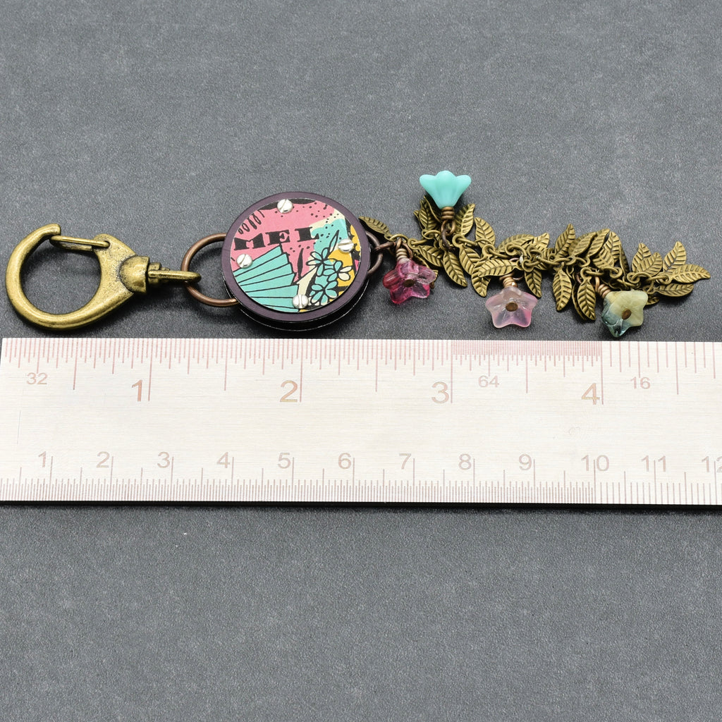 Decorative handbag charm with floral elements on a gray background with a ruler for scale.