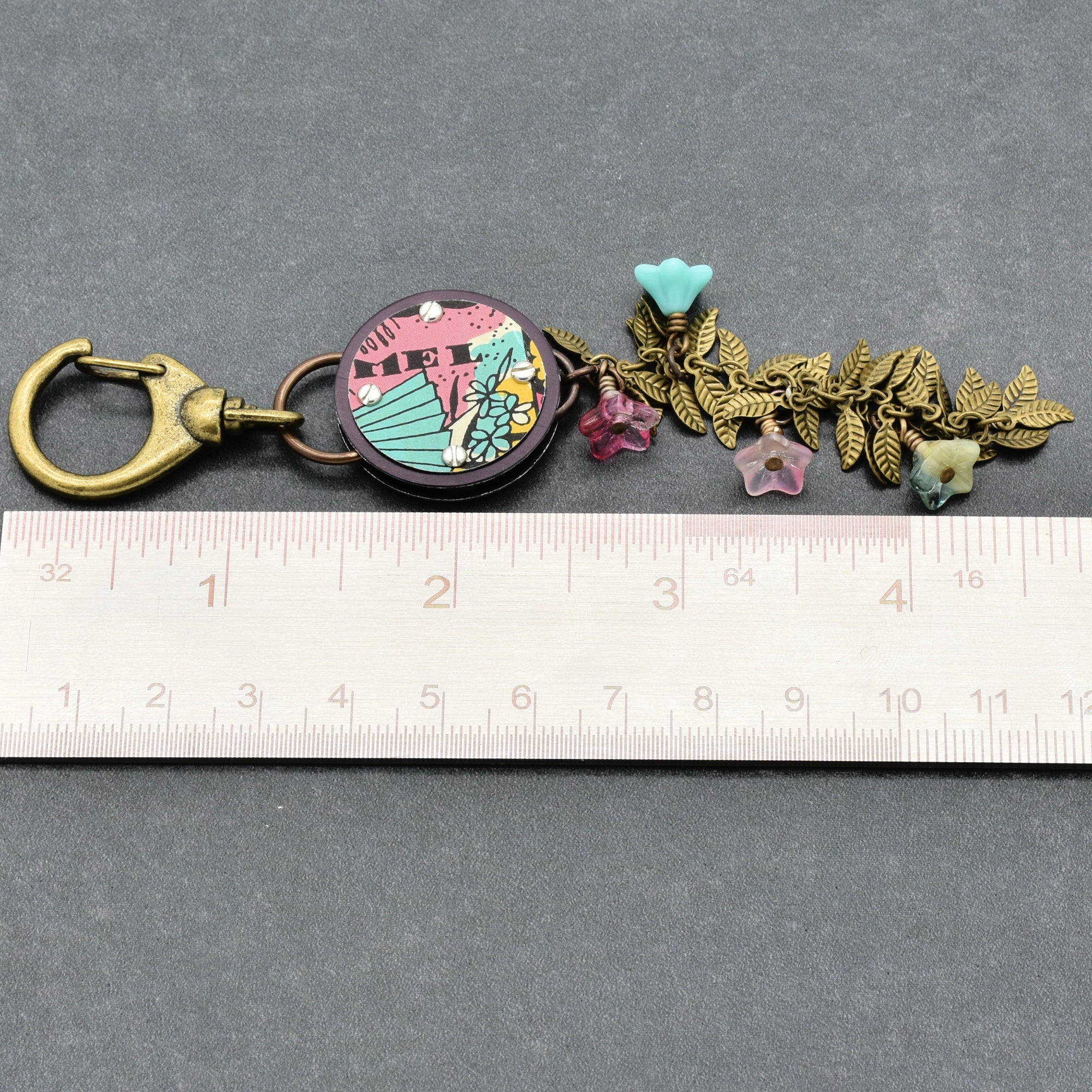 Decorative handbag charm with floral elements on a gray background with a ruler for scale.