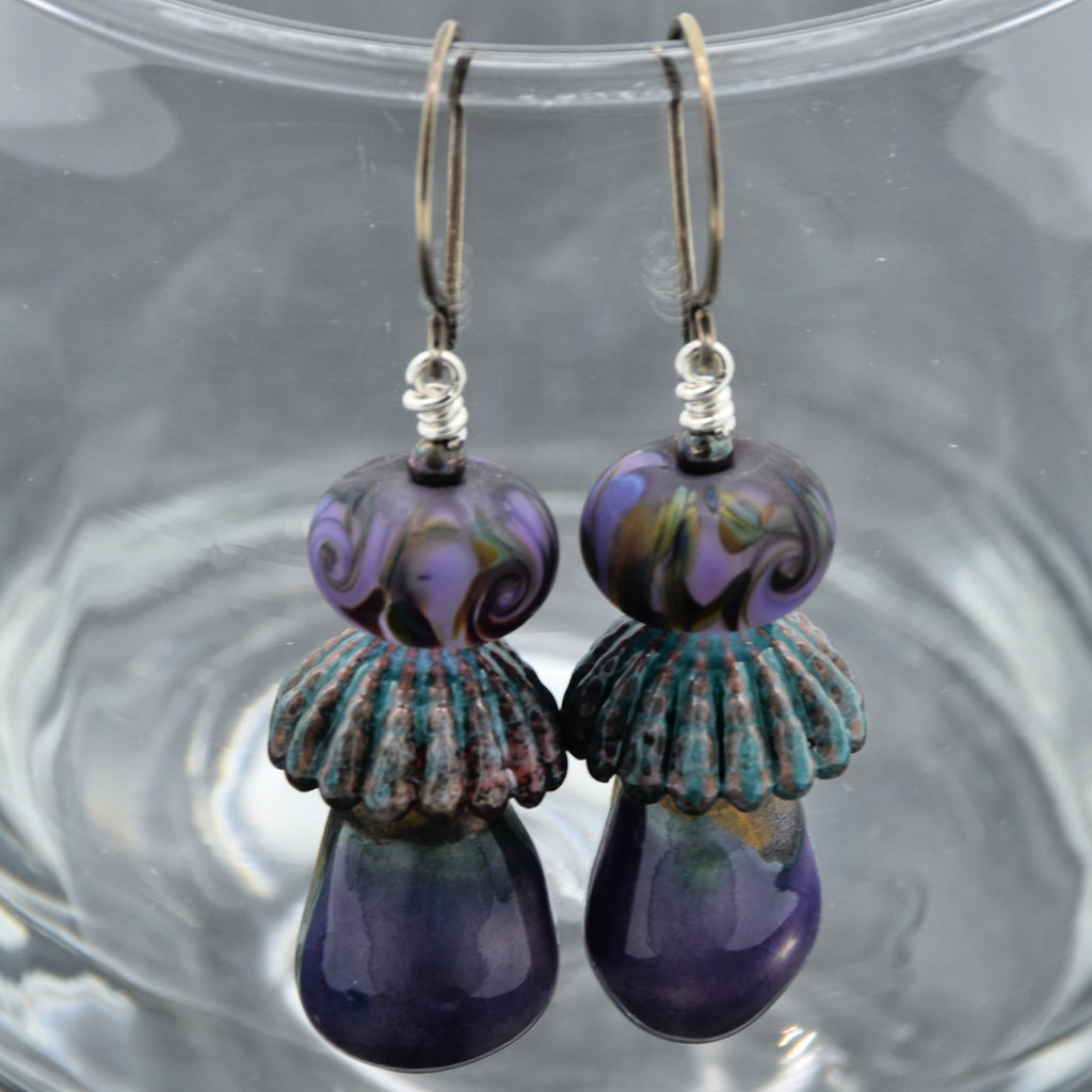 Pair of purple and teal lampwork glass and ceramic earrings on a clear glass surface