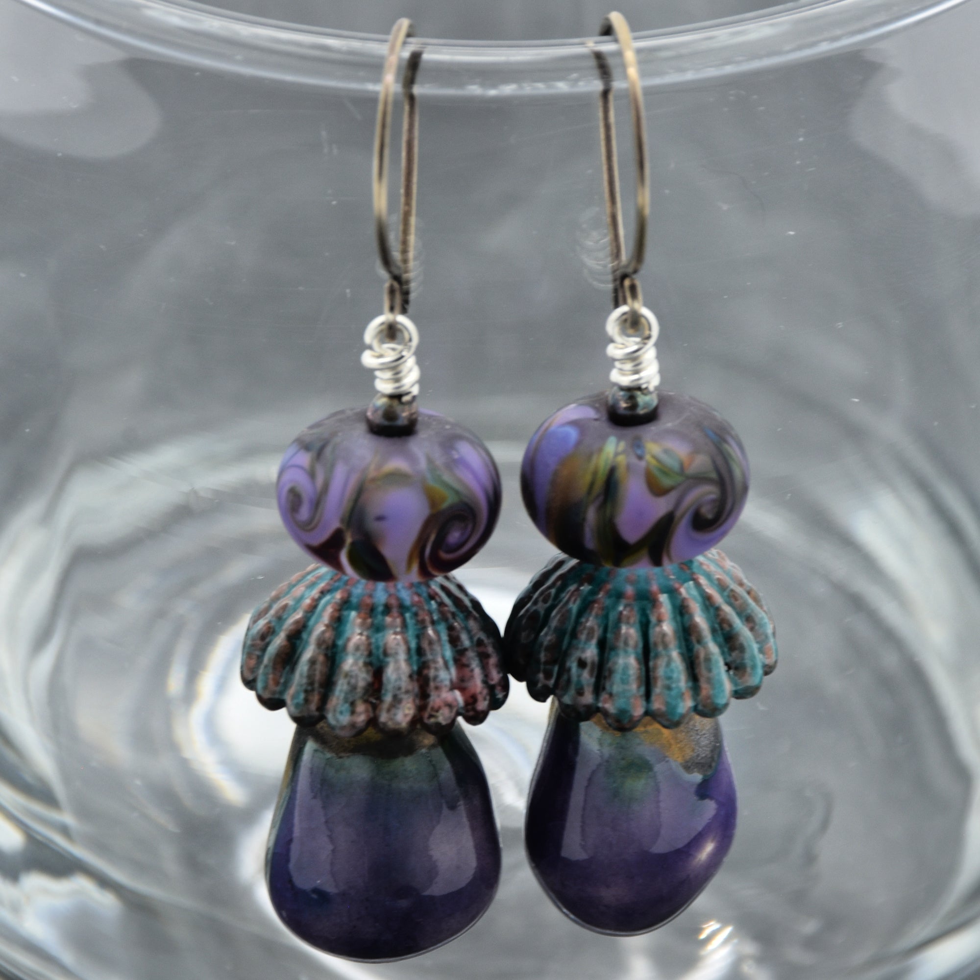 Pair of purple and teal lampwork glass and ceramic earrings on a clear glass surface