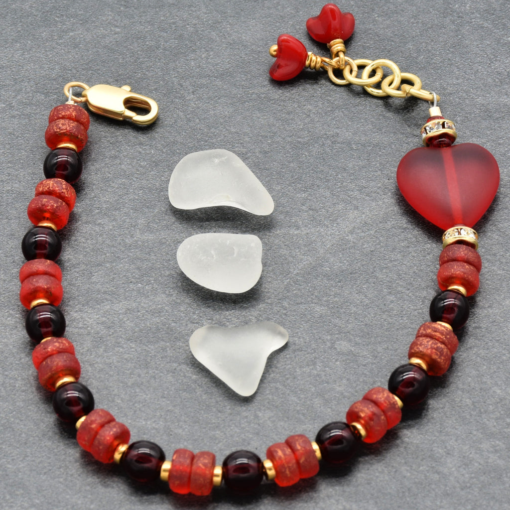 Bracelet with red and gold beads and heart-shaped charms on a gray surface with three pieces of sea glass.