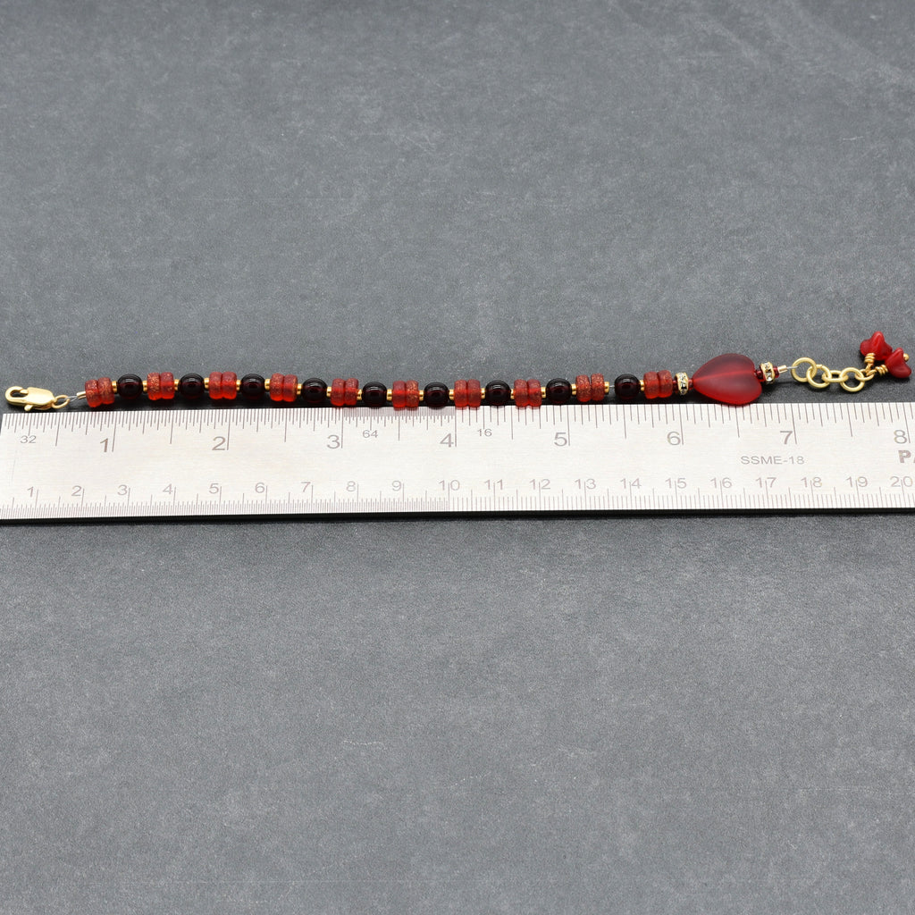 Beaded bracelet with red and gold accents on a gray surface, measured against a ruler.