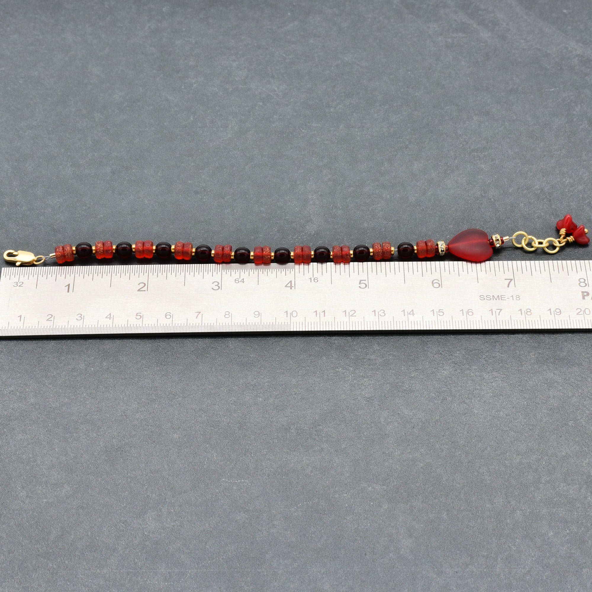 Beaded bracelet with red and gold accents on a gray surface, measured against a ruler.