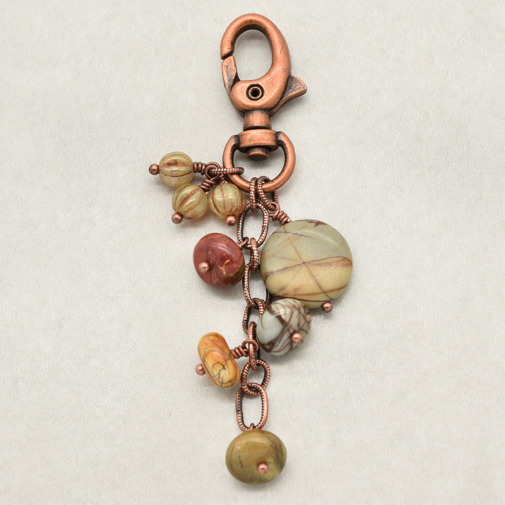 A handmade copper keychain featuring a lobster clasp, a ring, and dangling beads and charms made from natural stone beads in earthy green, brown, and orange hues, all wrapped in copper wire on a light beige background - made with love in Colorado by Ruby Acorn.