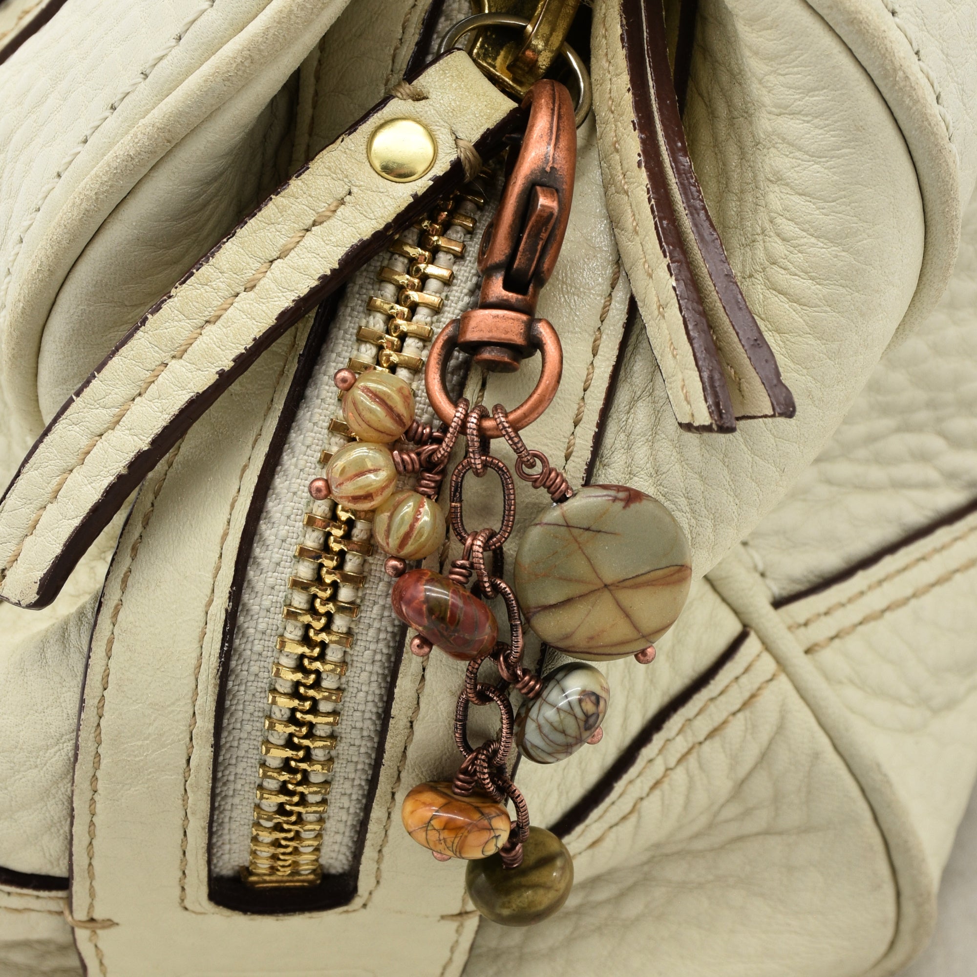 Close-up of a cream-colored leather bag featuring a decorative copper wire charm with natural stone beads and a gold-tone zipper pull - made with love in Colorado by Ruby Acorn.
