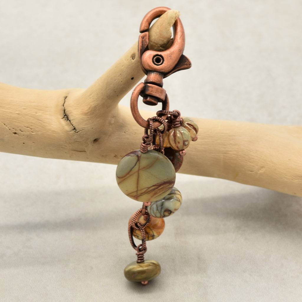 A handmade bohemian-style keychain or bag charm featuring a copper lobster clasp, a round marbled stone bead, and smaller accent beads wrapped in copper wire, displayed on a light wood branch against a neutral gray background - made with love in Colorado by Ruby Acorn.