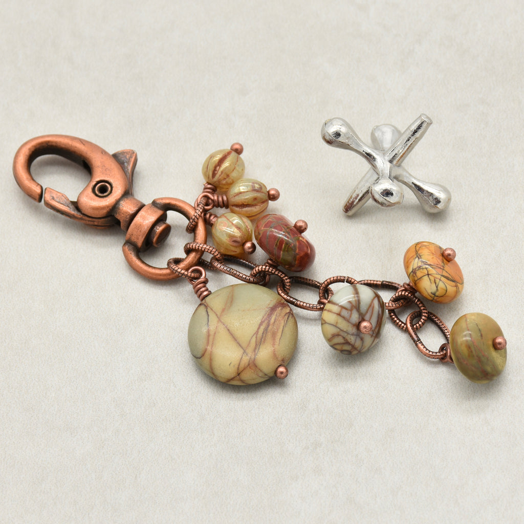 A handmade jewelry charm or keychain featuring a copper-toned lobster clasp, wire-wrapped copper chains, and multiple oval beads with a natural stone pattern in beige, brown, and olive green. A separate silver-toned metal piece is also shown on a textured light beige background - made with love in Colorado by Ruby Acorn.