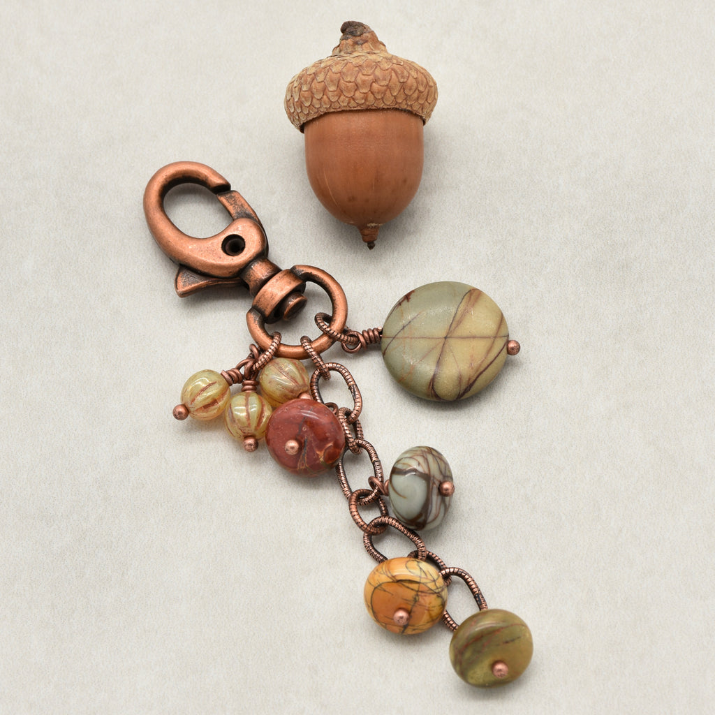A handmade keychain featuring a copper-toned lobster clasp, and multiple dangling round beads in earthy stone colors, displayed on a neutral textured background next to an acorn - made with love in Colorado by Ruby Acorn.