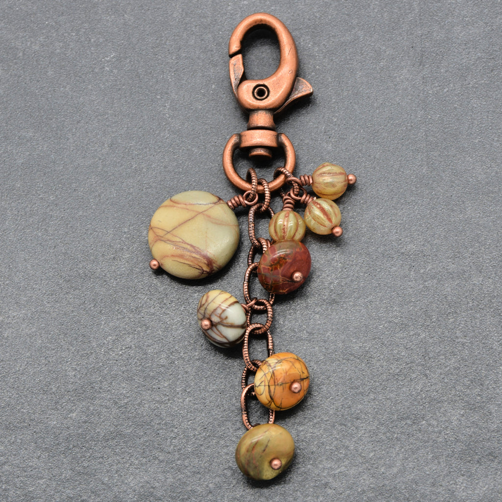 A handmade keychain featuring a copper-colored metal lobster clasp, ring, and chain, adorned with multiple round beads made from natural jasper stones in earthy green, brown, and yellow hues, set against a textured gray background - made with love in Colorado by Ruby Acorn.