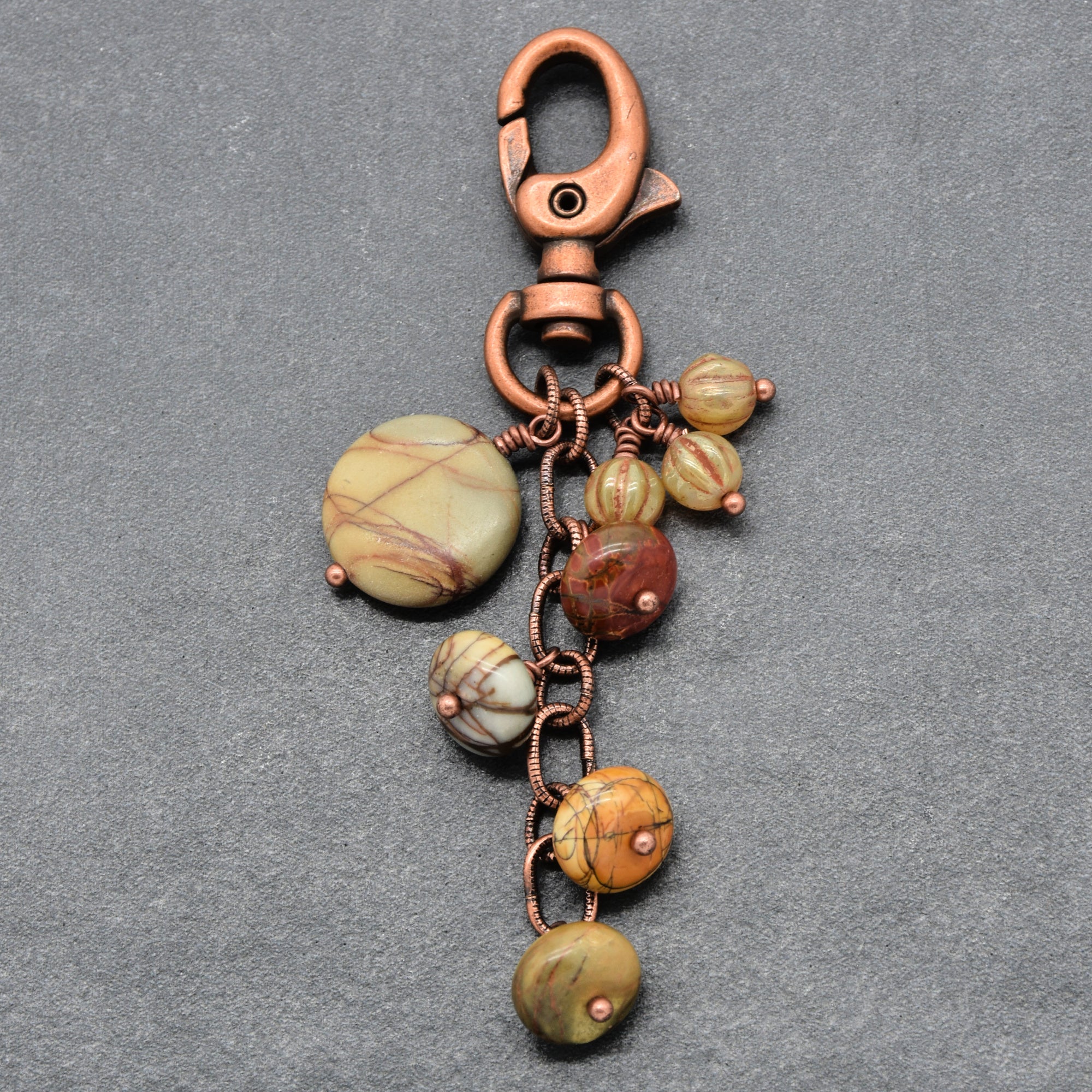 A handmade keychain featuring a copper-colored metal lobster clasp, ring, and chain, adorned with multiple round beads made from natural jasper stones in earthy green, brown, and yellow hues, set against a textured gray background - made with love in Colorado by Ruby Acorn.