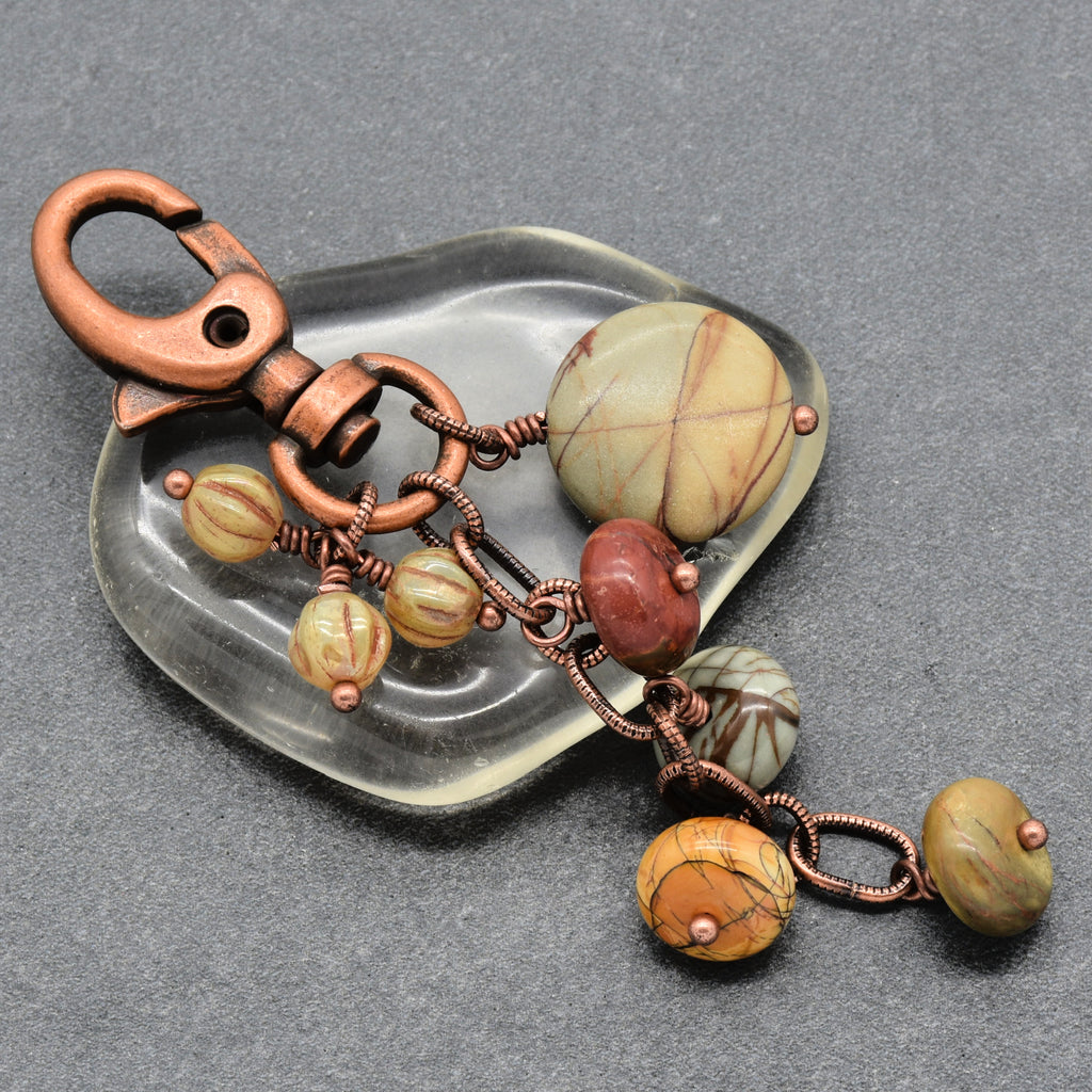A handmade jewelry keychain featuring a copper-toned lobster clasp and chain, adorned with textured stone beads in earthy yellow, brown, and reddish tones, displayed on a clear glass dish over a gray background - made with love in Colorado by Ruby Acorn.