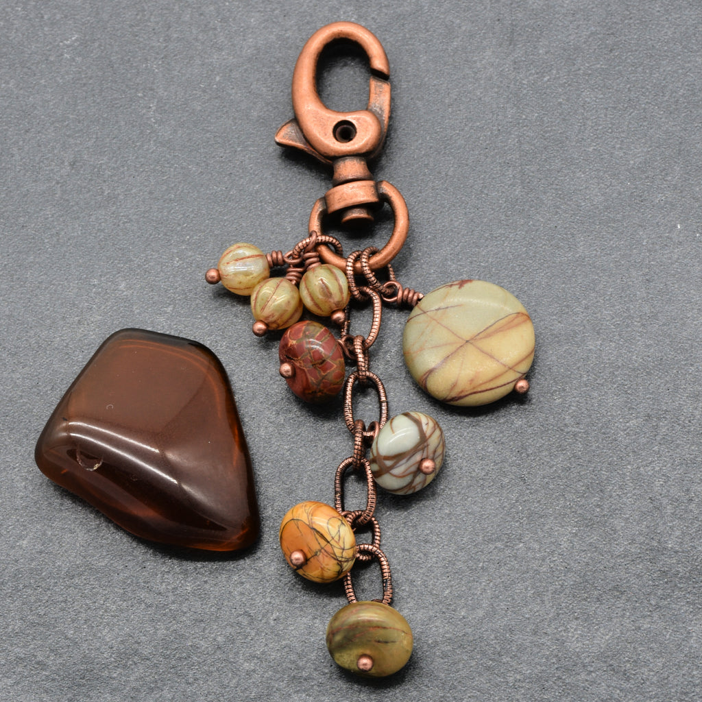 A handmade keychain featuring a copper-colored lobster clasp, a short chain with links, and several beads and pendants made from natural stone in shades of, brown, red and cream, displayed on a textured gray background made with love in Colorado by Ruby Acorn.