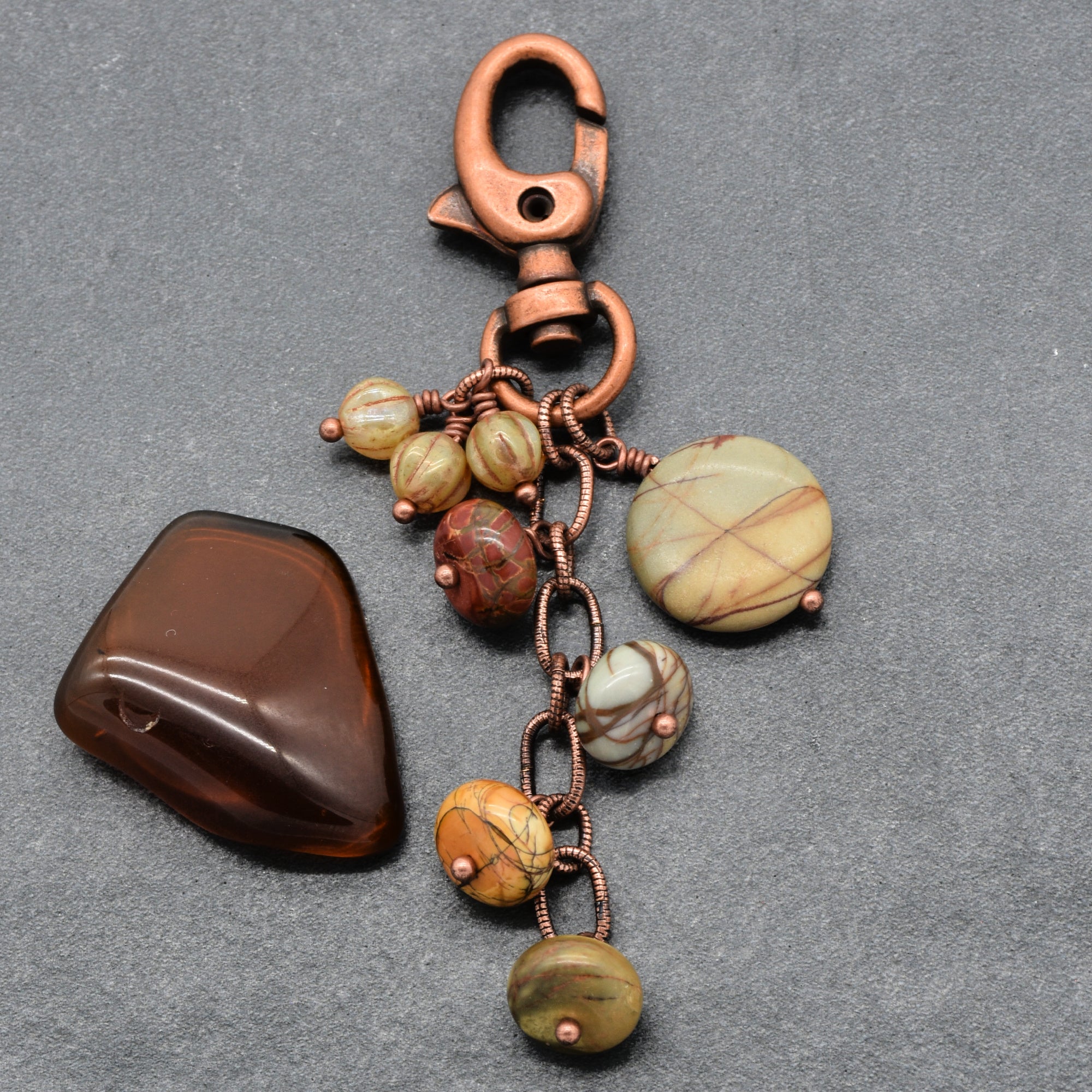 A handmade keychain featuring a copper-colored lobster clasp, a short chain with links, and several beads and pendants made from natural stone in shades of, brown, red and cream, displayed on a textured gray background made with love in Colorado by Ruby Acorn.