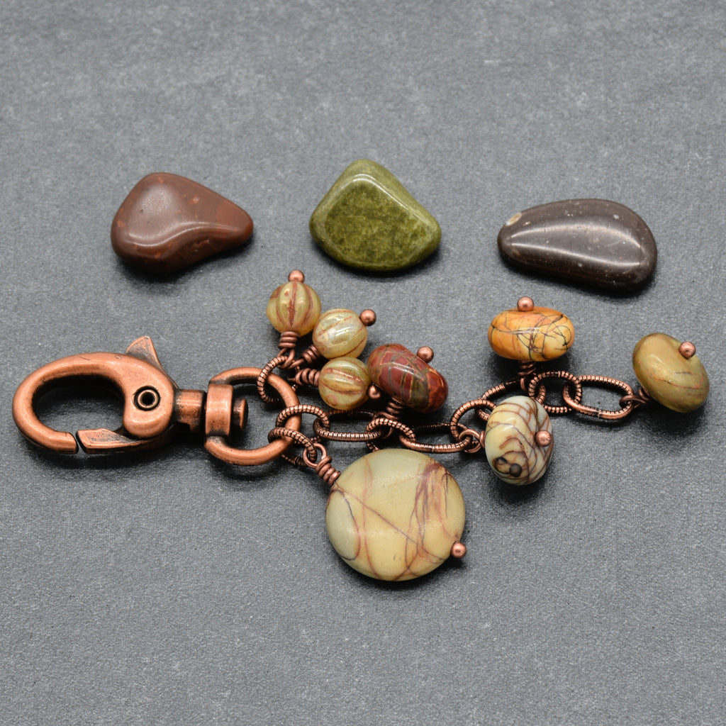 A handmade keychain or bag charm made of copper wire and beads, featuring a large round beige and brown stone pendant, smaller patterned beads, and a copper clasp, displayed on a gray background with three decorative stones - made with love in Colorado by Ruby Acorn.
