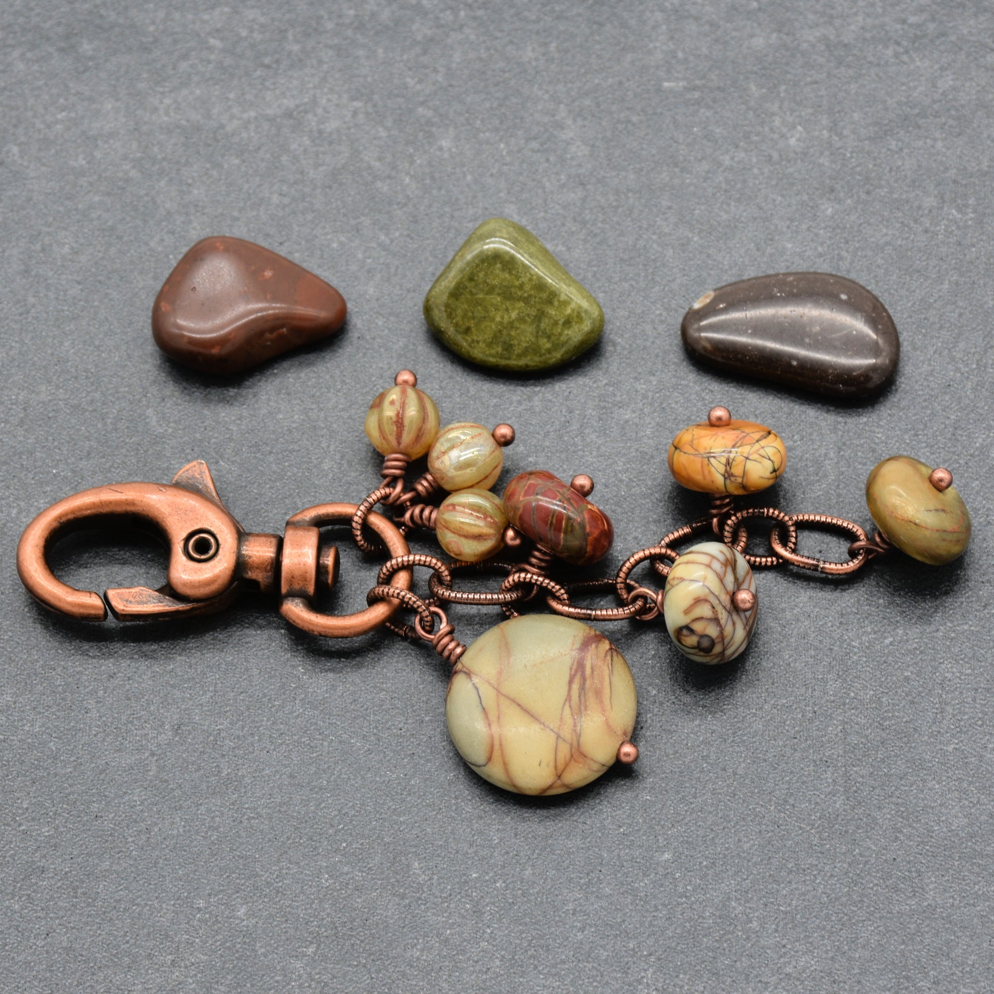 A handmade keychain or bag charm made of copper wire and beads, featuring a large round beige and brown stone pendant, smaller patterned beads, and a copper clasp, displayed on a gray background with three decorative stones - made with love in Colorado by Ruby Acorn.