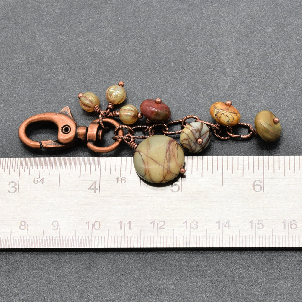 A handmade keychain or bag charm featuring natural stone beads in earthy green, brown, and reddish tones, wrapped in copper wire with a copper clasp, shown next to a metal ruler for scale on a dark gray background - made with love in Colorado by Ruby Acorn.