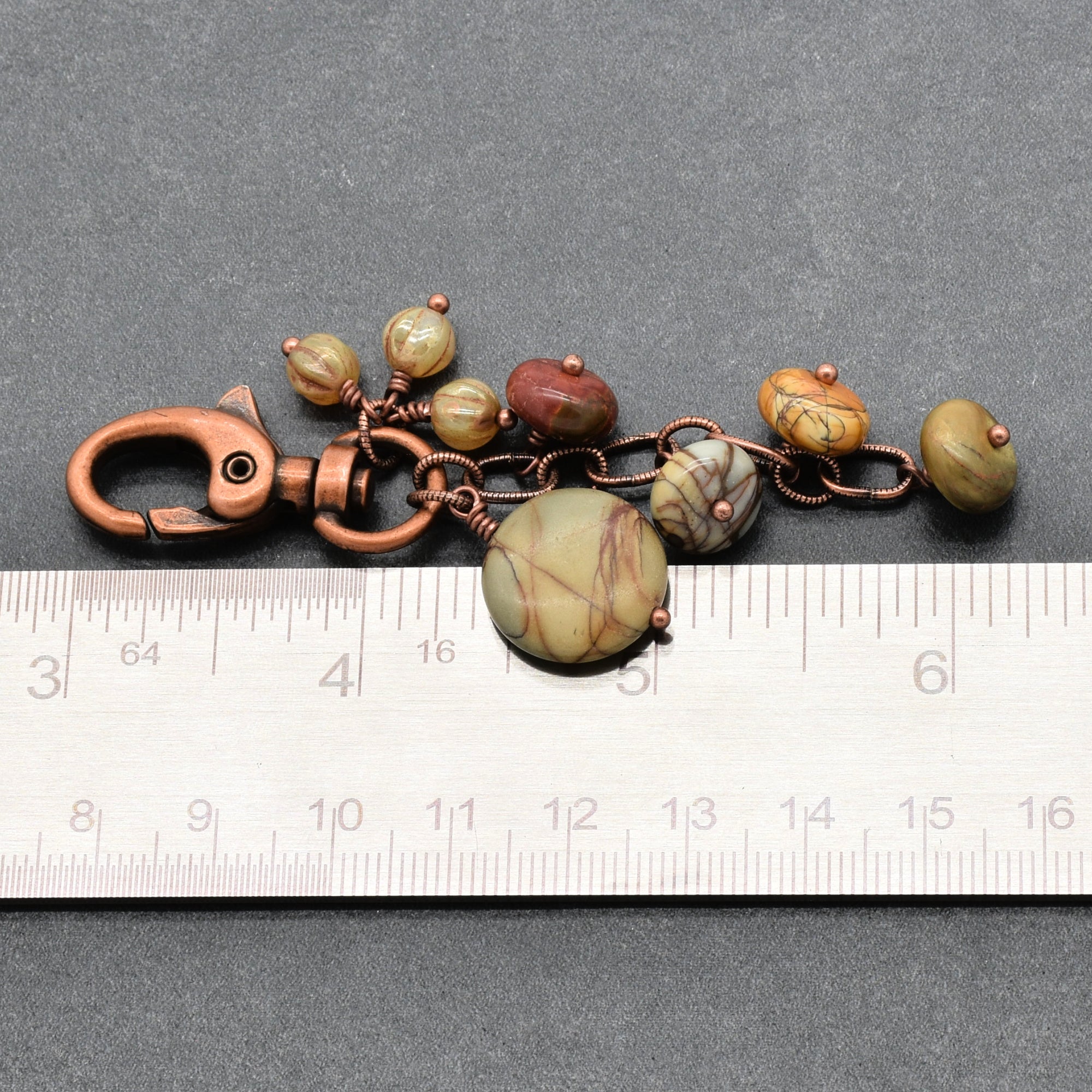 A handmade keychain or bag charm featuring natural stone beads in earthy green, brown, and reddish tones, wrapped in copper wire with a copper clasp, shown next to a metal ruler for scale on a dark gray background - made with love in Colorado by Ruby Acorn.