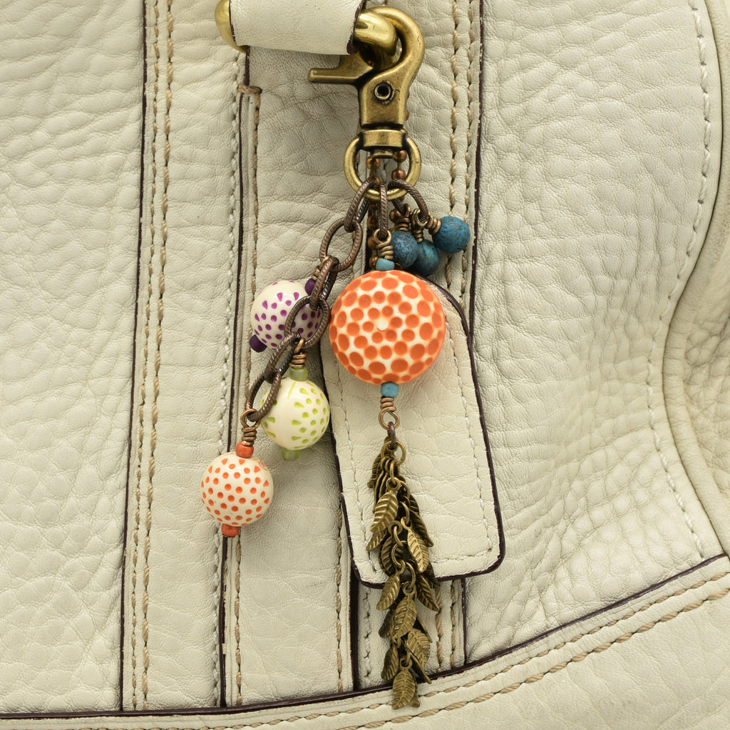 A close-up of a cream-colored textured leather bag featuring a decorative keychain or bag charm attached near the zipper. The charm includes a brass-tone clasp, small blue beads, patterned vintage Lucite beads in orange, green, and purple, and a dangling bronze-toned leaf tassel - made with love in Colorado by Ruby Acorn.