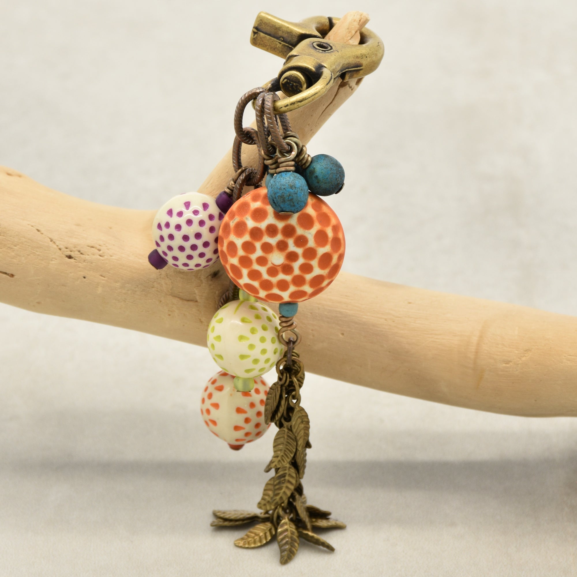 A bohemian-style beaded charm with a brass-toned clasp and dangling charms, displayed on a light wood branch. The bracelet features a prominent orange beaded sphere, smaller patterned beads in purple, green, and orange, and blue beads, finished with a leaf chain - made with love in Colorado by Ruby Acorn.