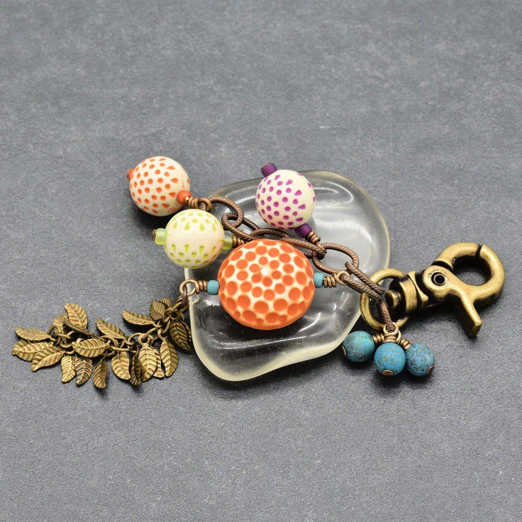 A decorative keychain or bag charm with an antique brass clasp, featuring patterned vintage Lucite beads in orange, yellow, and purple, turquoise Czech glass beads, antique brass falling leaves chain  on a gray background - made with love in Colorado by Ruby Acorn.