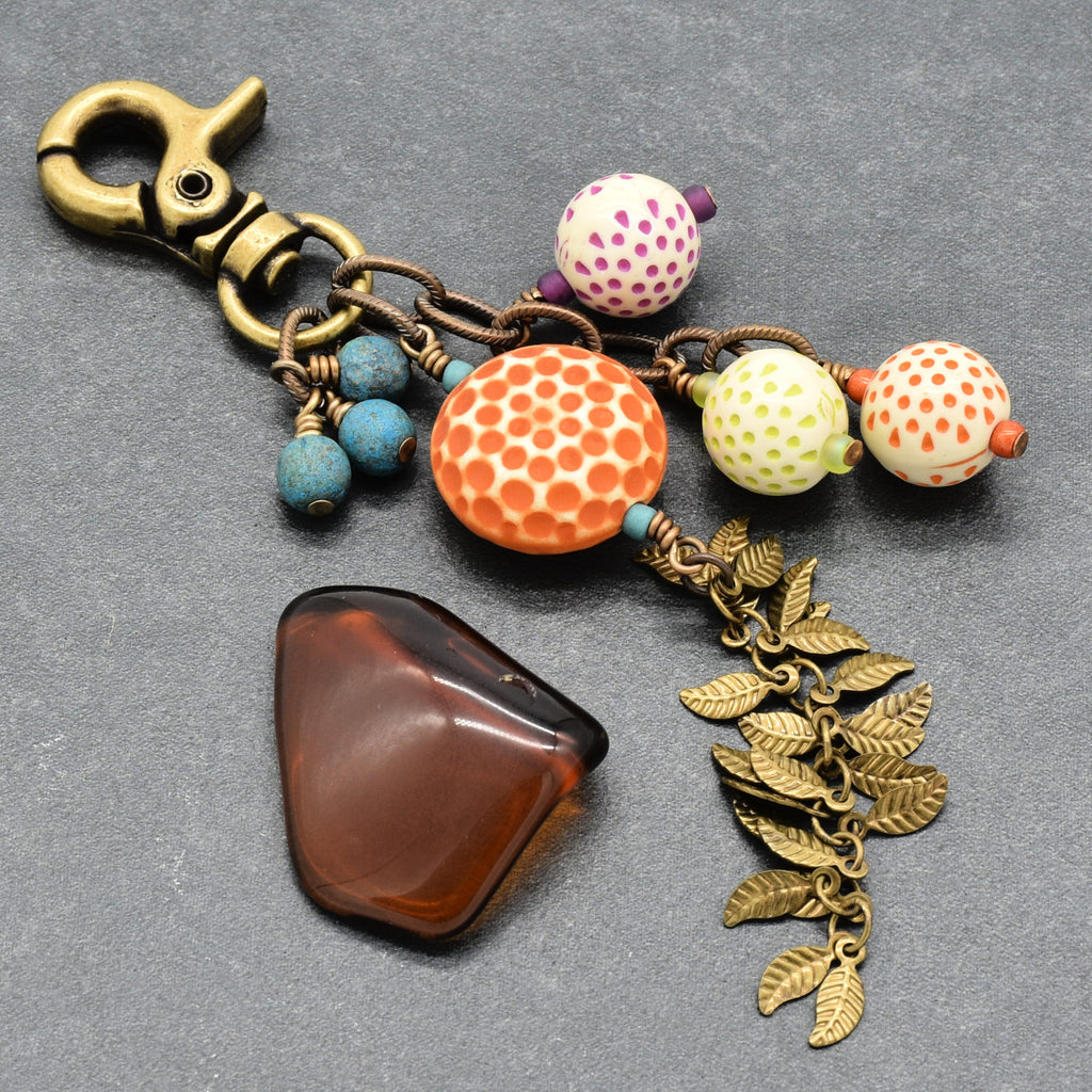 A handmade beaded keychain or bag charm featuring a brass clasp, vintage Lucite beads, Czech glass, and antique brass-toned falling leaves chain, displayed on a textured gray background - handmade with love in Colorado by Ruby Acorn.