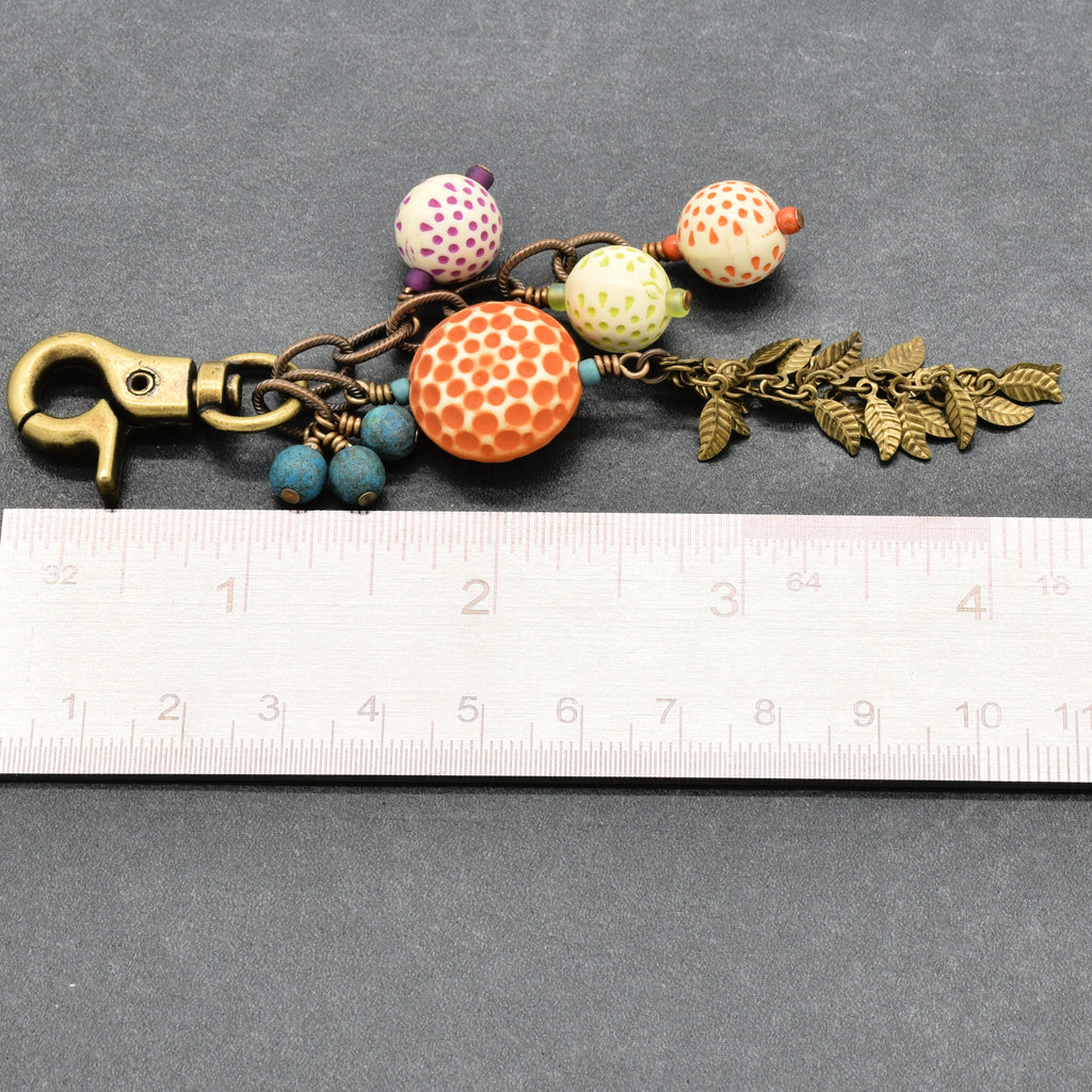 A handmade keychain or bag charm with a brass clasp, patterned beads, a large orange honeycomb bead, blue beads, and antique brass falling leaves chain, shown next to a white ruler on a gray background - made with love in Colorado by Ruby Acorn.