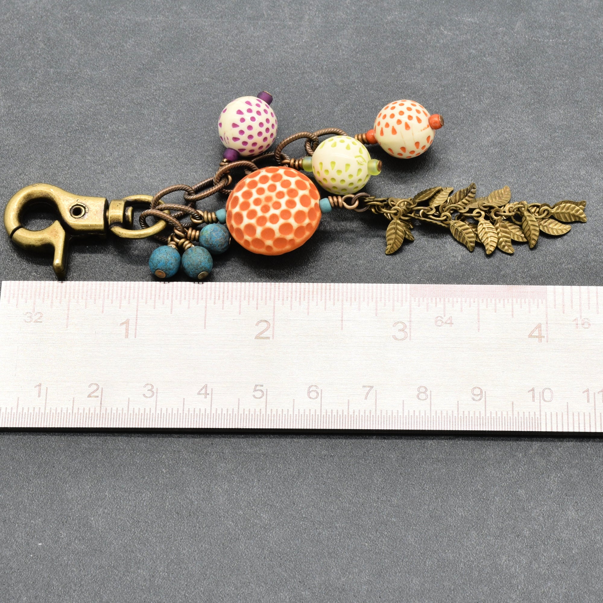 A handmade keychain or bag charm with a brass clasp, patterned beads, a large orange honeycomb bead, blue beads, and antique brass falling leaves chain, shown next to a white ruler on a gray background - made with love in Colorado by Ruby Acorn.