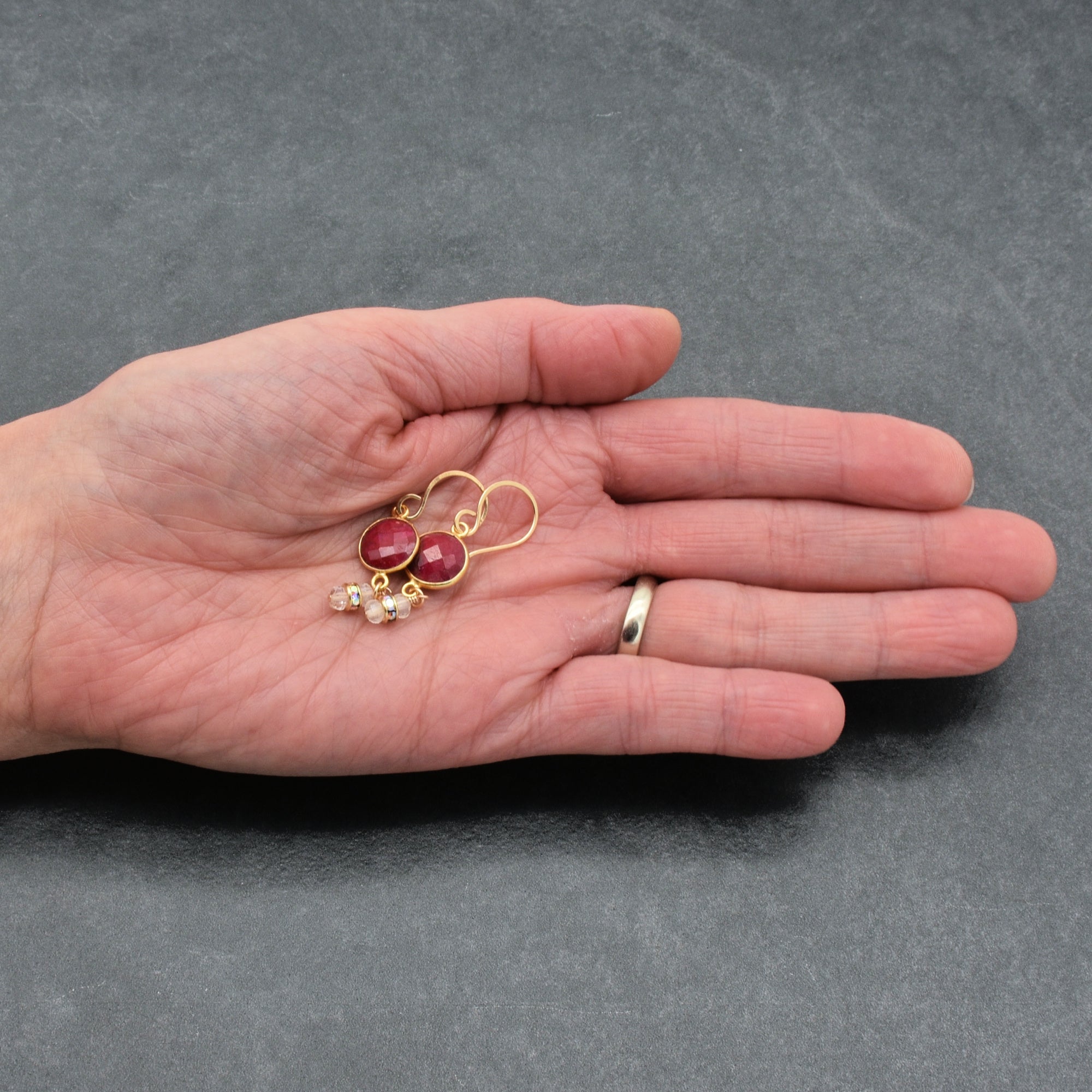 Gold earrings with red stones held in a hand against a gray background