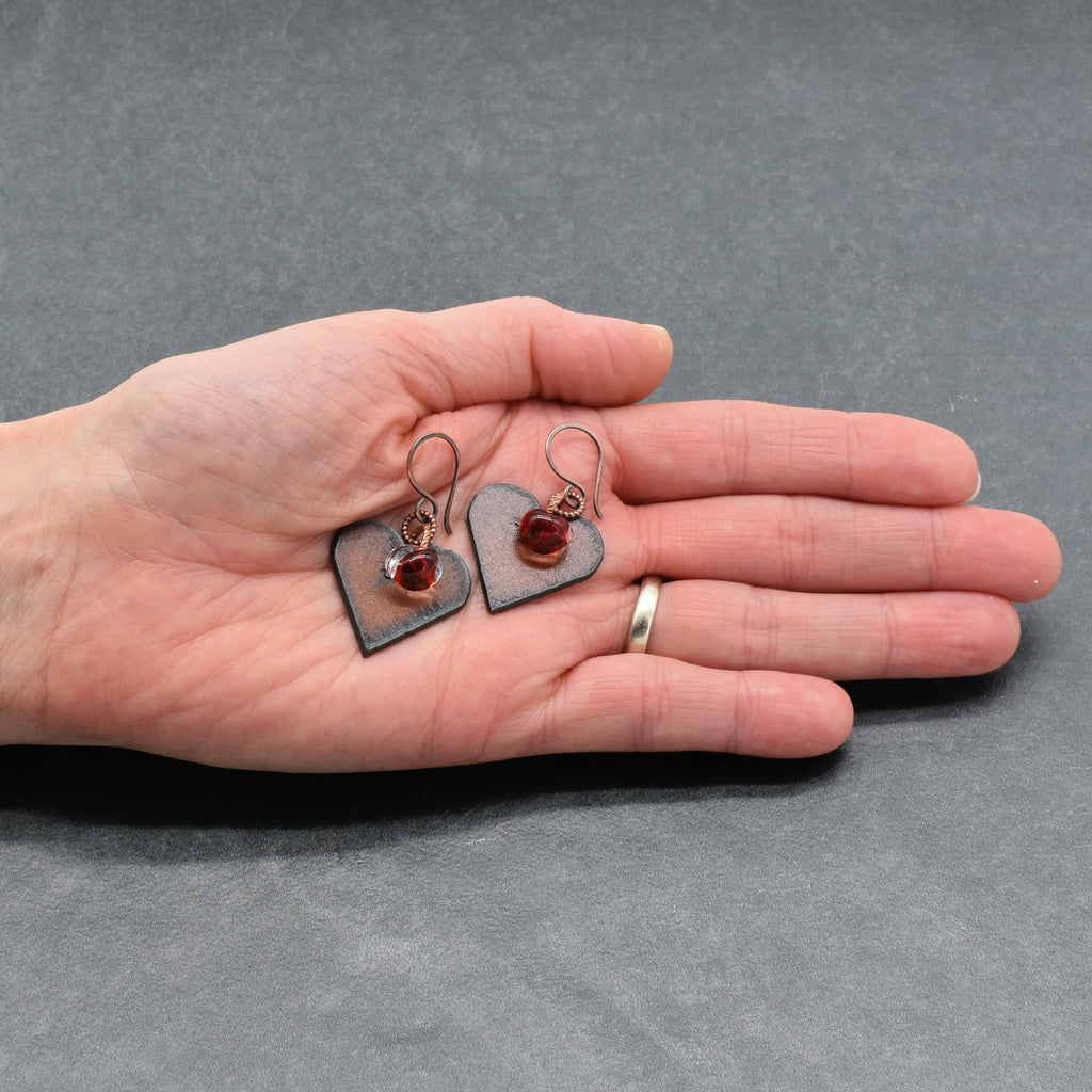 Heart-shaped earrings with red charms held in a hand against a gray background