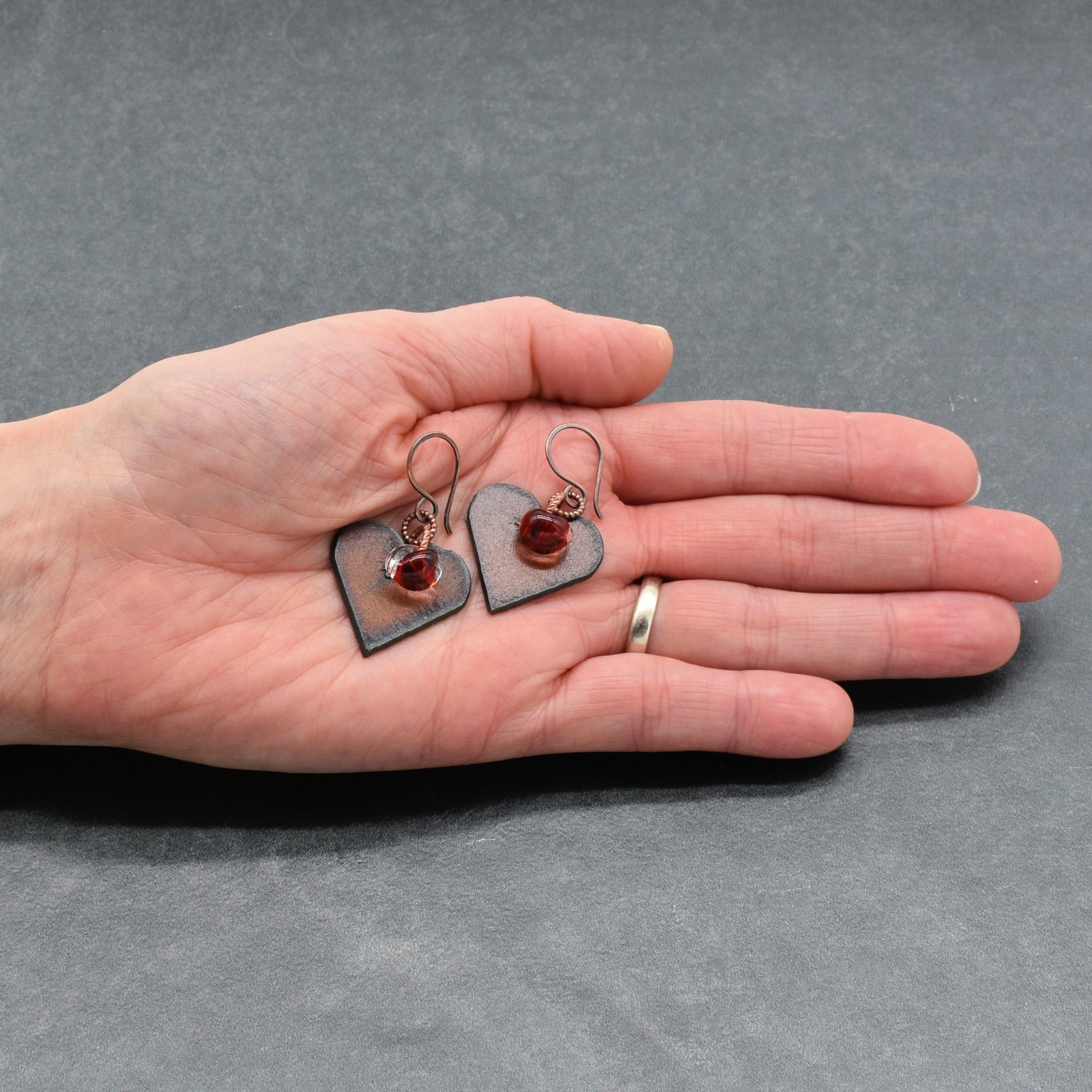 Heart-shaped earrings with red charms held in a hand against a gray background