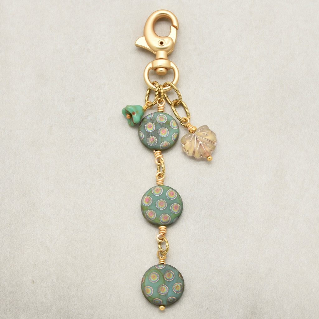 A handmade keychain or bag charm featuring three round sage green glass beads with gold and pink polka dots, a small green flower bead, a clear iridescent leaf charm, and a gold lobster clasp on a light gray background - made with love in Colorado by Ruby Acorn.