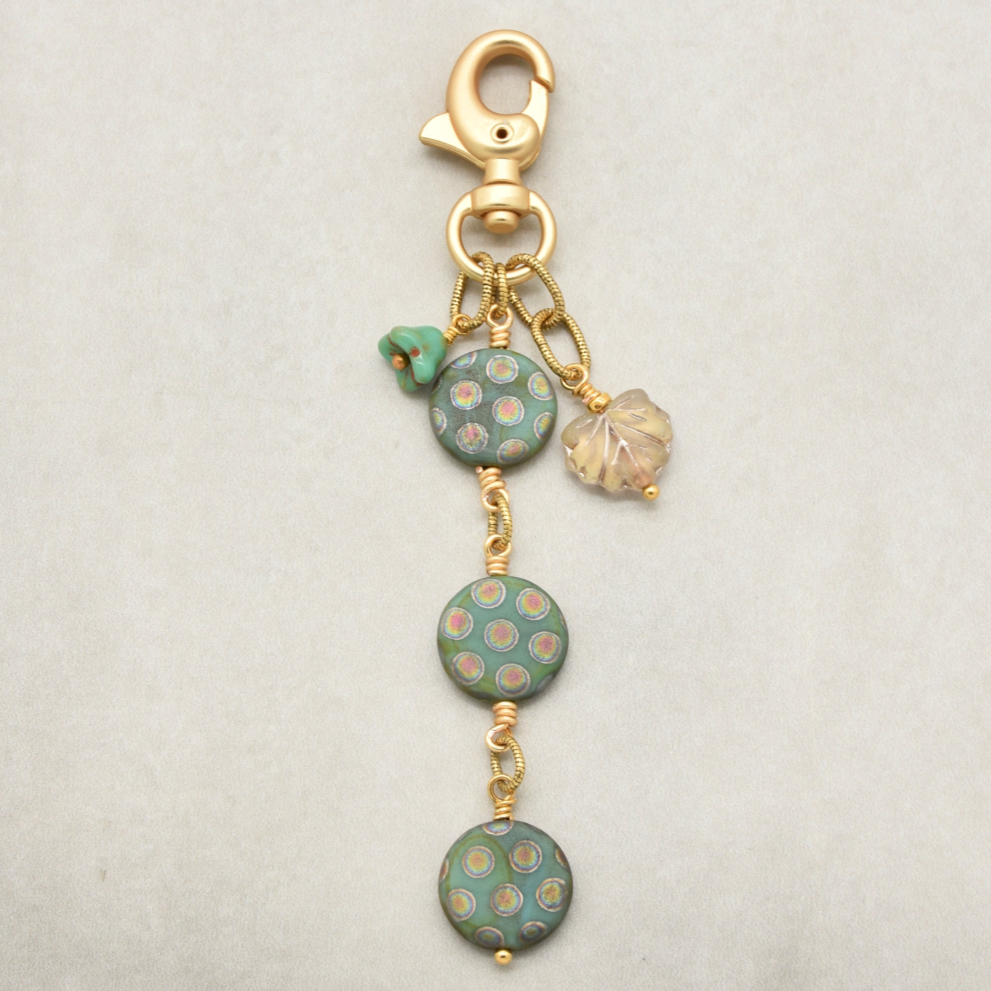 A handmade keychain or bag charm featuring three round sage green glass beads with gold and pink polka dots, a small green flower bead, a clear iridescent leaf charm, and a gold lobster clasp on a light gray background - made with love in Colorado by Ruby Acorn.
