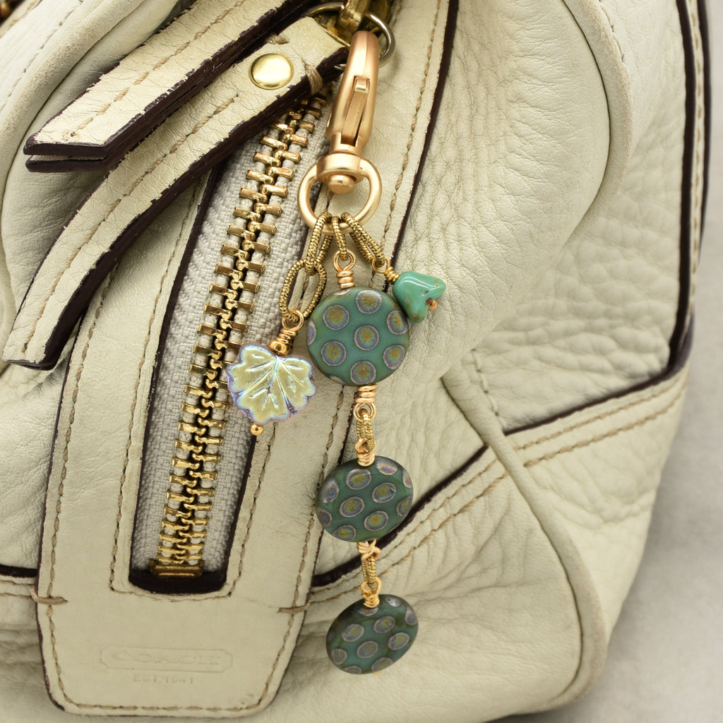 Close-up of a handbag in light beige leather with a gold zipper, featuring a decorative keychain charm with green patterned discs, a flower bead, and a small iridescent leaf charm - made with love in Colorado by Ruby Acorn.