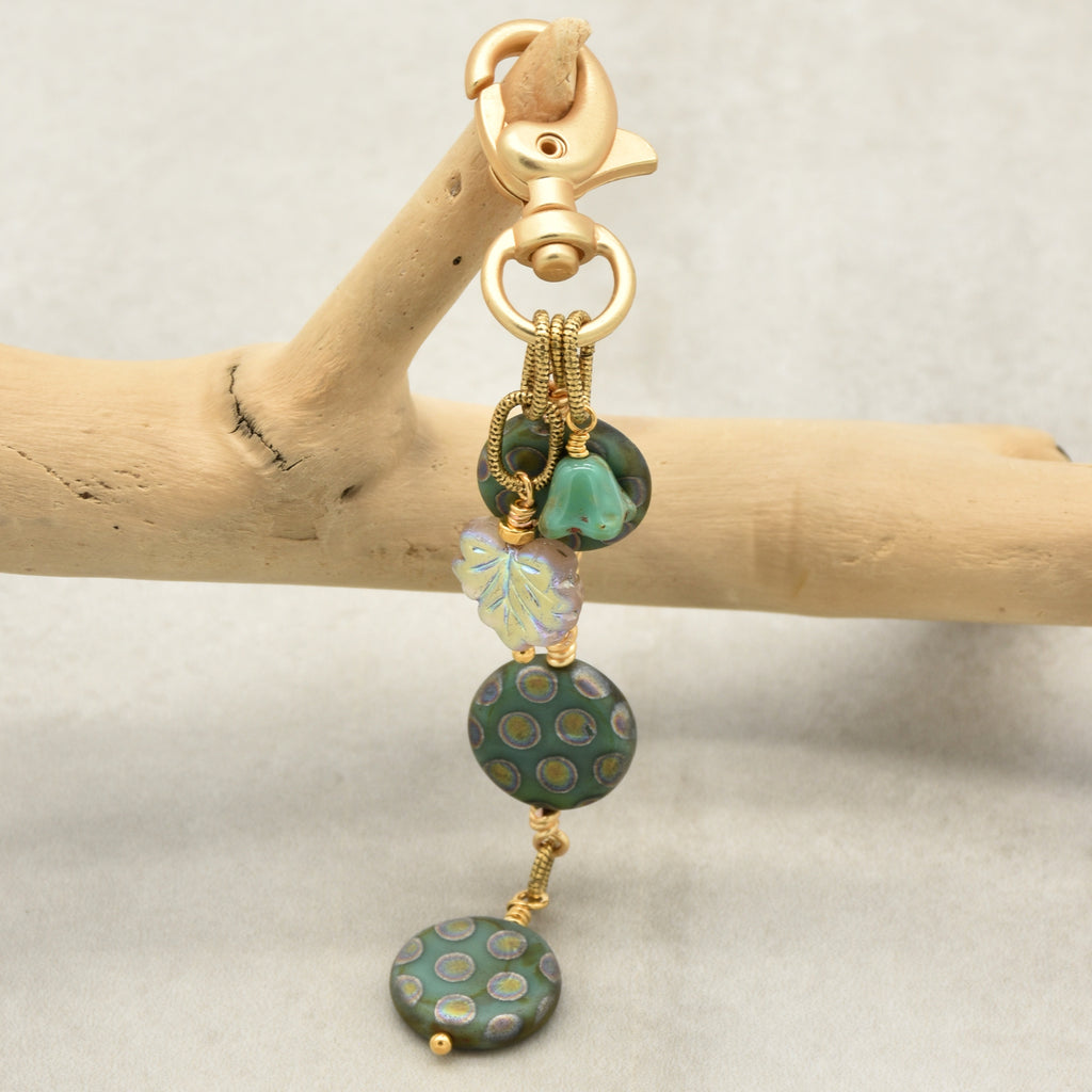 A handmade beaded keychain or bag charm with a gold-tone lobster clasp, featuring green patterned beads, a turquoise green flower, and a light iridescent leaf bead, displayed on a light wood branch against a neutral gray background - made with love in Colorado by Ruby Acorn.