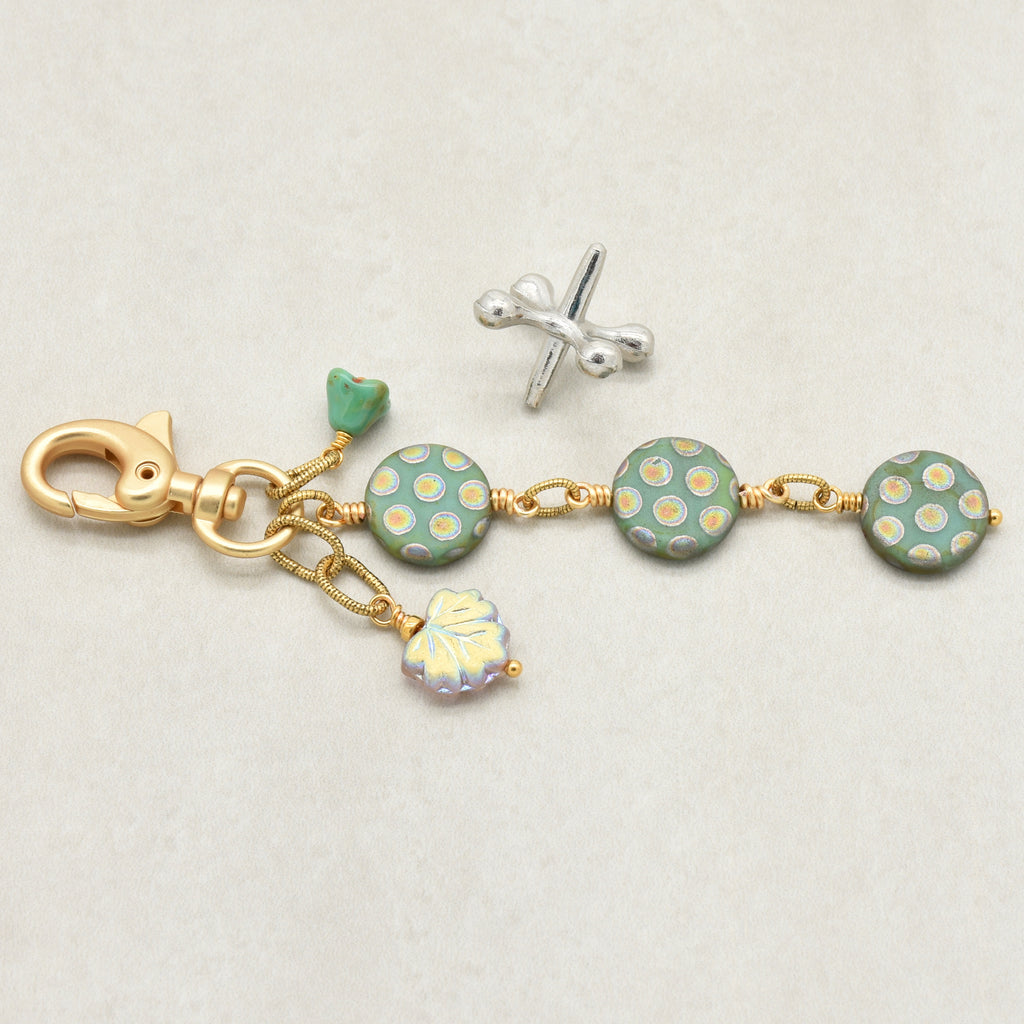 A handmade keychain or bag charm with a gold-tone lobster clasp, three round sage green beads with gold polka dots, a small green flower charm, and a dangling iridescent leaf charm, shown on a light beige background - made with love in Colorado by Ruby Acorn.