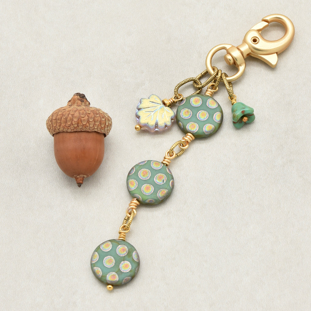 An autumn-themed keychain charm featuring three round sage green beads with yellow and white polka dots, a leaf charm, and a small green flower, all attached to a gold clasp on a light gray background - made with love in Colorado by Ruby Acorn.