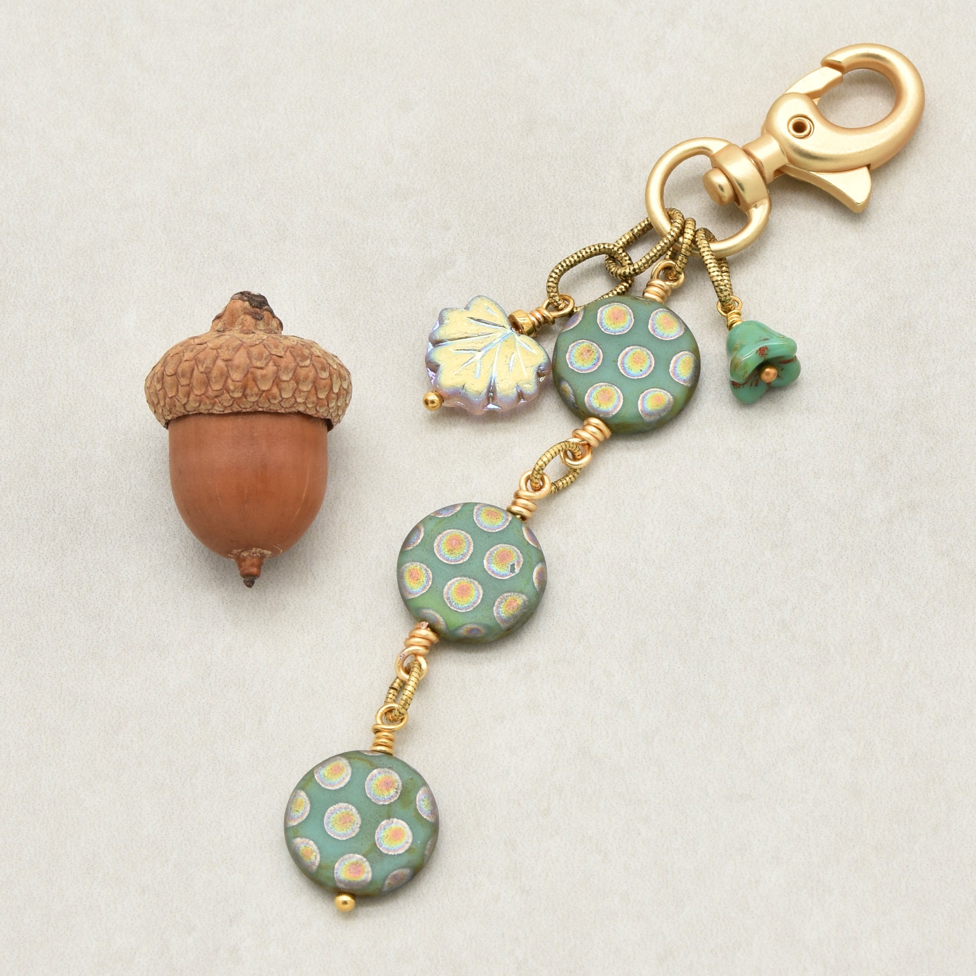 An autumn-themed keychain charm featuring three round sage green beads with yellow and white polka dots, a leaf charm, and a small green flower, all attached to a gold clasp on a light gray background - made with love in Colorado by Ruby Acorn.