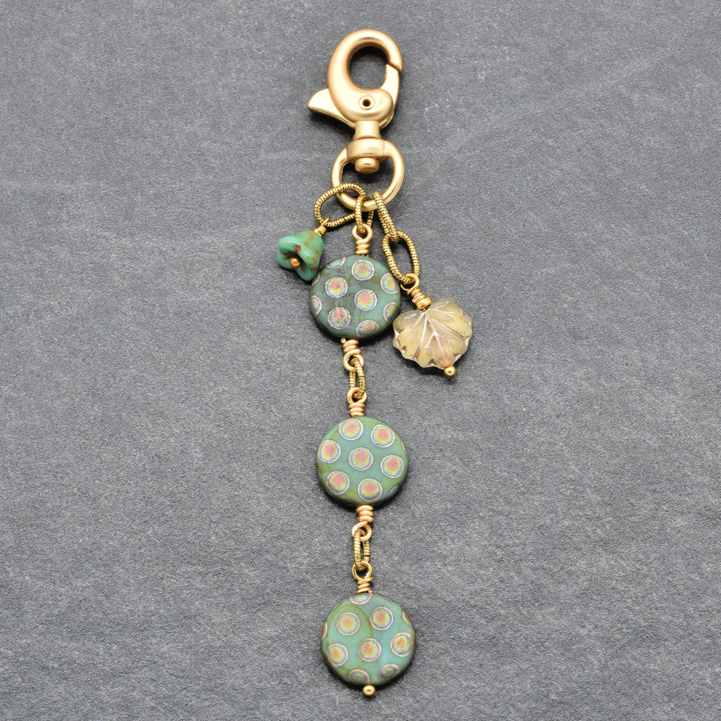 A vintage-style gold-tone keychain or bag charm with three round green beads, a small green bead, and a leaf charm, displayed on a textured gray background - made with love in Colorado by Ruby Acorn.
