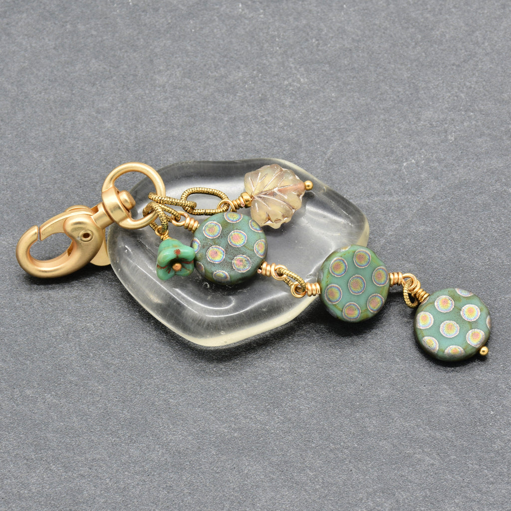 A handmade keychain or bag charm featuring green patterned glass beads, a small green flower bead, a clear leaf-shaped bead, and a gold-tone clasp, displayed on a gray textured background - made with love in Colorado by Ruby Acorn.