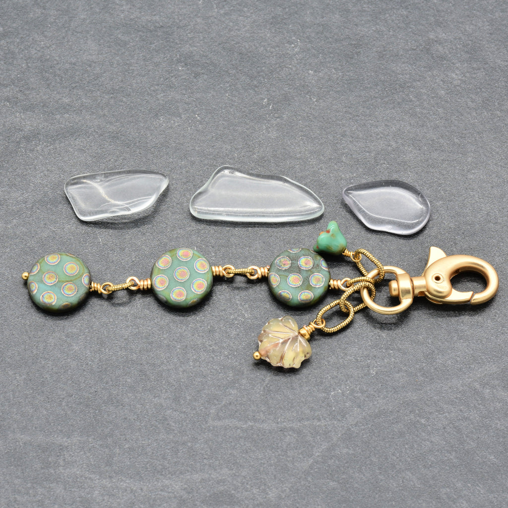 A handmade keychain or bag charm laid flat on a dark gray textured background. The charm features three round green beads with a gold polka-dot pattern, a small green flower charm, a gold leaf charm, and a gold lobster clasp - made with love in Colorado by Ruby Acorn.