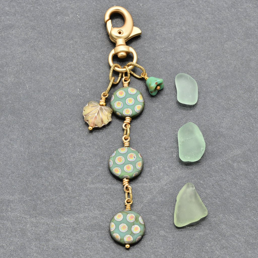 A handmade sage green keychain charm with three coin Czech glass beads in sage green, a small green bellflower Czech glass bead, a gold-toned glass leaf charm, and a gold lobster clasp, displayed next to three matching sea glass pieces on a gray background - made with love in Colorado by Ruby Acorn.