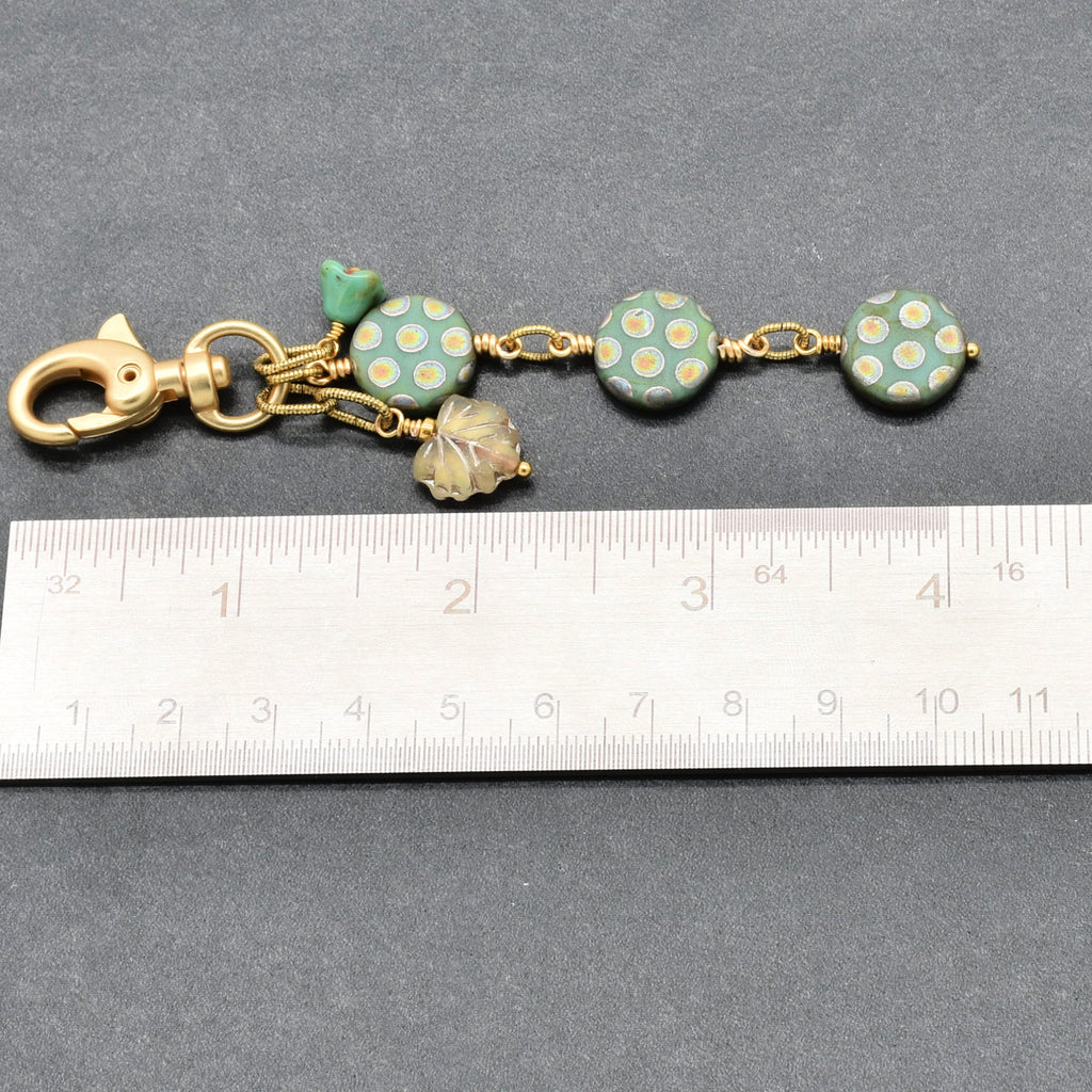 A handmade keychain or bag charm with a gold-tone clasp, featuring three round sage green beads with white polka dots, a small green flower bead, and a leaf charm, shown next to a metal ruler for scale on a gray background - made with love in Colorado by Ruby Acorn.