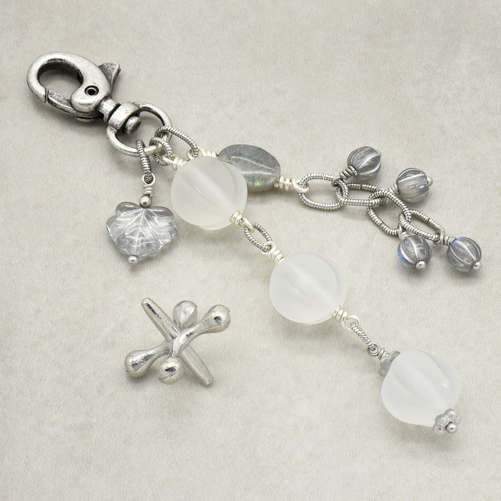 Snowfall Serenade handmade bag charm with three frosted white fluted Lucite beads, a labradorite oval, gray Czech glass beads, a clear glass maple leaf, and an antique-silver-finish swivel clasp on a light background.