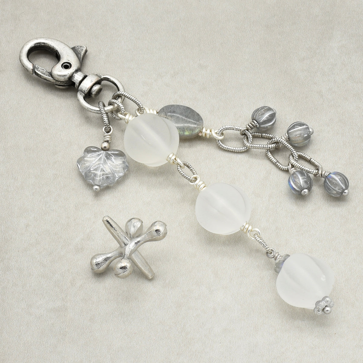 Snowfall Serenade handmade bag charm with three frosted white fluted Lucite beads, a labradorite oval, gray Czech glass beads, a clear glass maple leaf, and an antique-silver-finish swivel clasp on a light background.