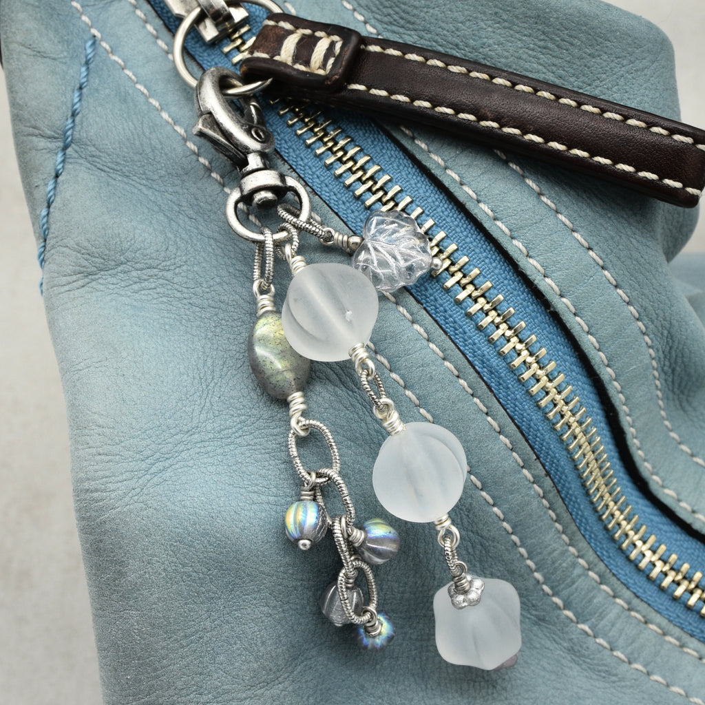 Snowfall Serenade handmade bag charm with three frosted white fluted Lucite beads, a labradorite oval, gray Czech glass beads, a clear glass maple leaf, and an antique-silver-finish swivel clasp on a light blue leather handbag.