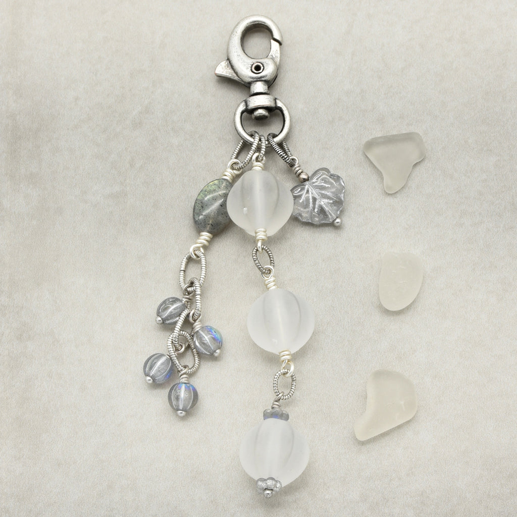 Snowfall Serenade handmade bag charm with three frosted white fluted Lucite beads, a labradorite oval, gray Czech glass beads, a clear glass maple leaf, and an antique-silver-finish swivel clasp on a light background.