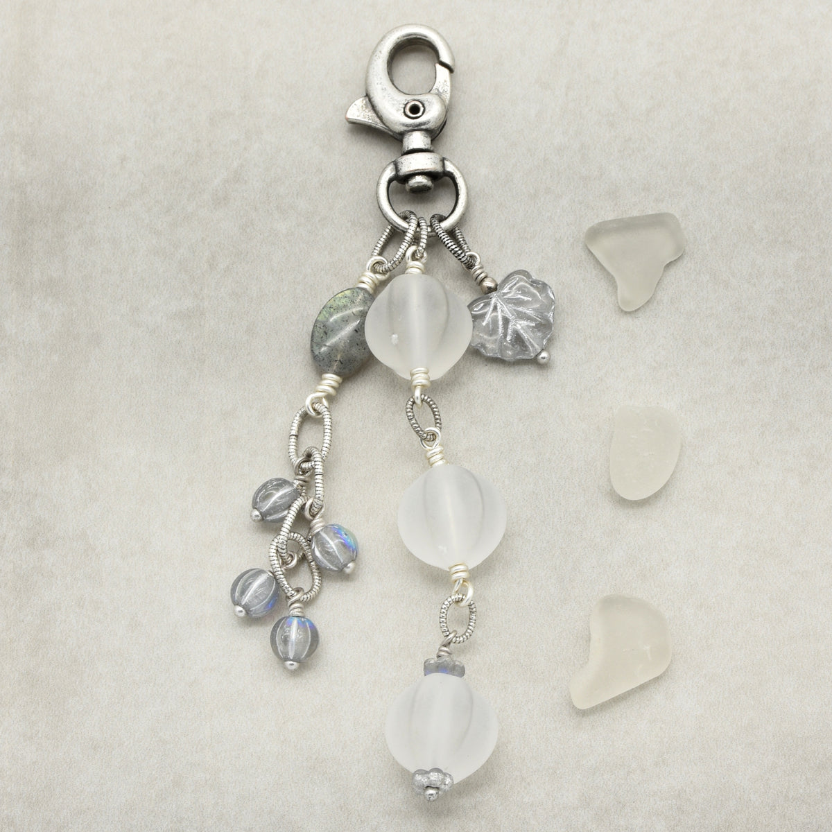 Snowfall Serenade handmade bag charm with three frosted white fluted Lucite beads, a labradorite oval, gray Czech glass beads, a clear glass maple leaf, and an antique-silver-finish swivel clasp on a light background.