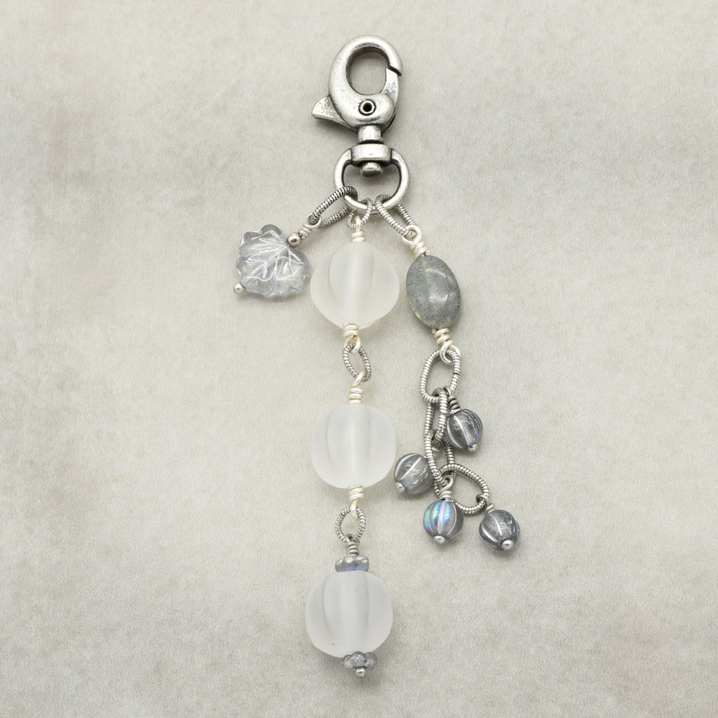 Snowfall Serenade handmade bag charm with three frosted white fluted Lucite beads, a labradorite oval, gray Czech glass beads, a clear glass maple leaf, and an antique-silver-finish swivel clasp on a light background.