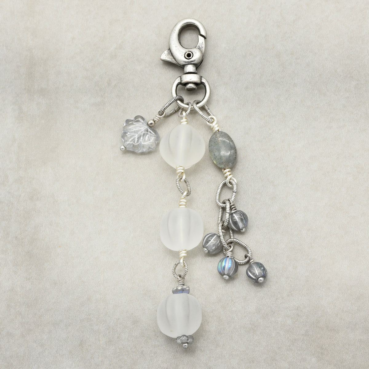 Snowfall Serenade handmade bag charm with three frosted white fluted Lucite beads, a labradorite oval, gray Czech glass beads, a clear glass maple leaf, and an antique-silver-finish swivel clasp on a light background.